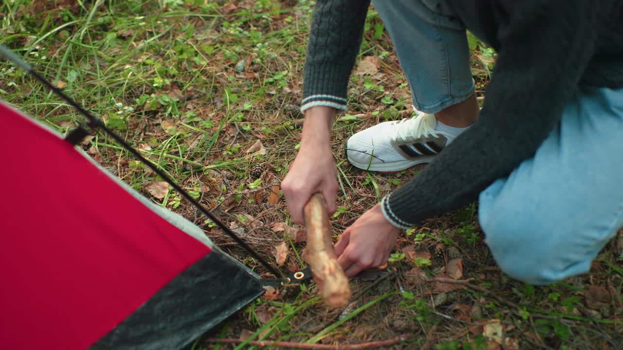 close up of boy squatting beside red tent hammering metal peg into forest floor using wooden stick during camping trip wearing sweater and jeans surrounded by grass dry leaves and branches outdoors