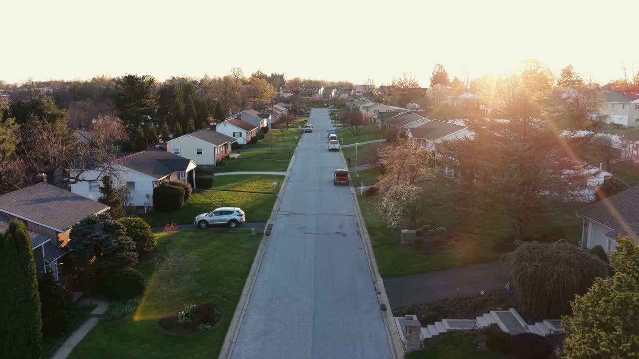 Vista aérea de drone subindo sobre um bairro suburbano americano ao nascer do sol, mostrando casas, telhados, jardins e ruas tranquilas banhadas por uma luz dourada e quente em uma área residencial pacífica dos EUA
