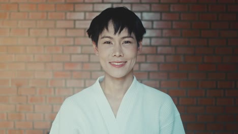 woman in a robe, smiling at the camera in front of a brick wall