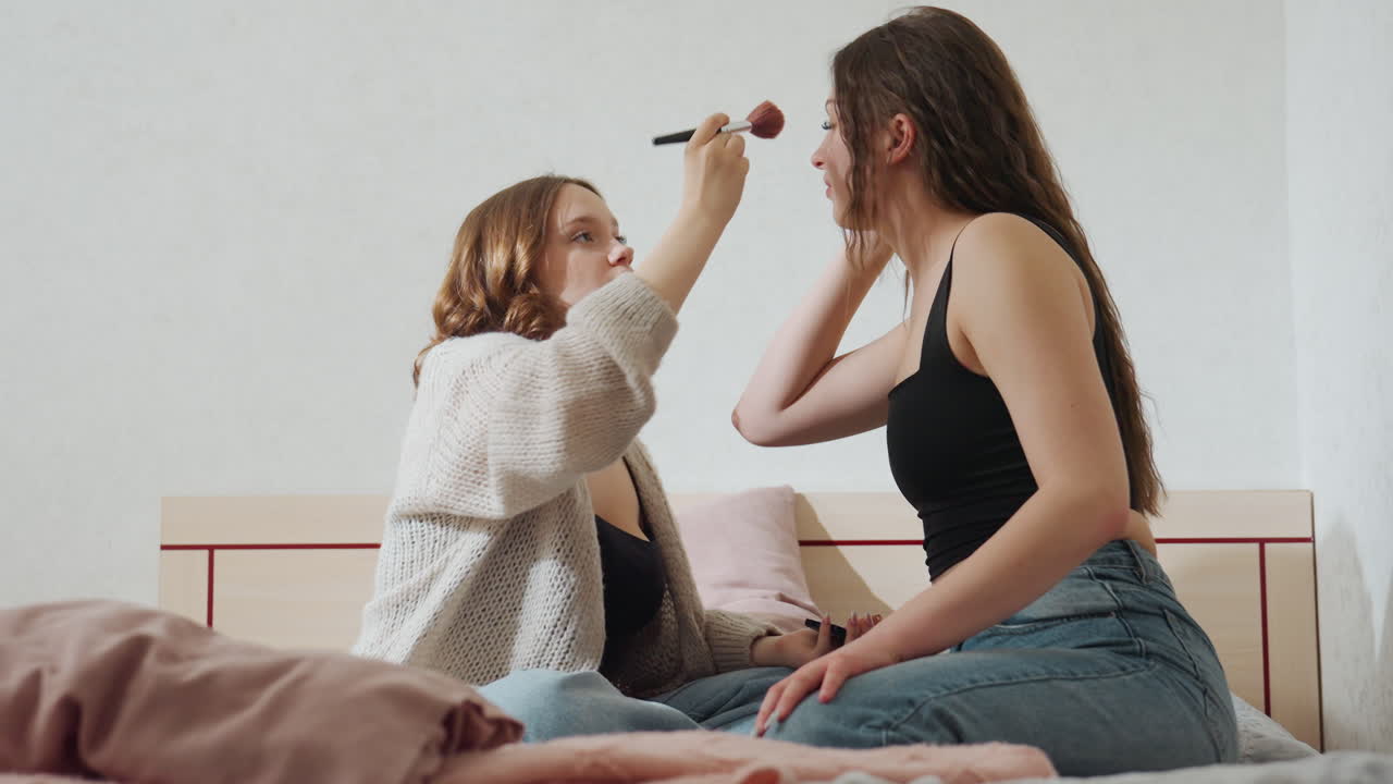 Caucasian Friends Applying Makeup On Bed, Cozy Morning Scene With One Friend Using Brush To Blend Blush On Other Cheek, Soft Natural Light, Casual Jeans And Sweater, Warm Smiles And Laughter, Candid