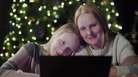 mom and daughter are looking at the laptop screen together. sitting in front of the blurred lights of the christmas tree