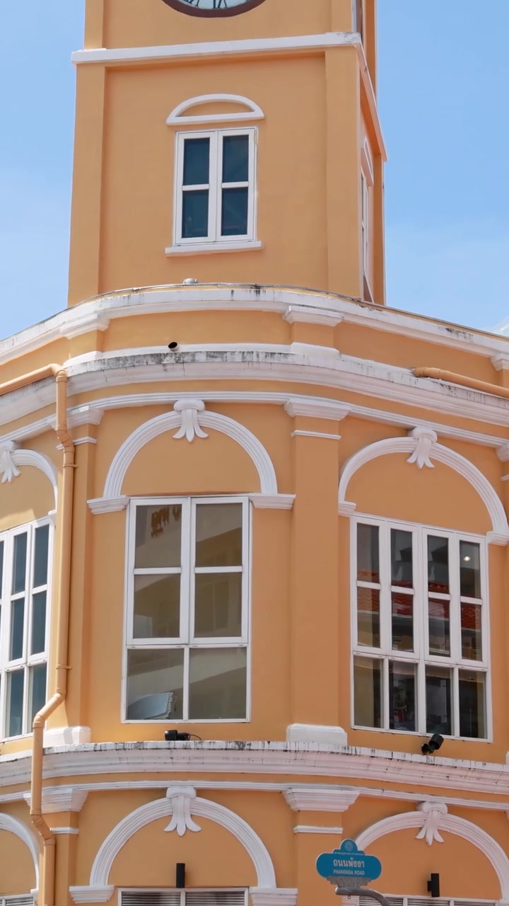 Uma torre do relógio na Cidade Velha de Phuket, com veículos em trânsito, sob luz diurna intensa, exibindo arquitetura colonial e vida urbana.