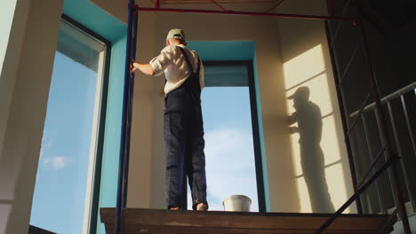 a builder in a protective helmet primes the wall, stands high on scaffolding