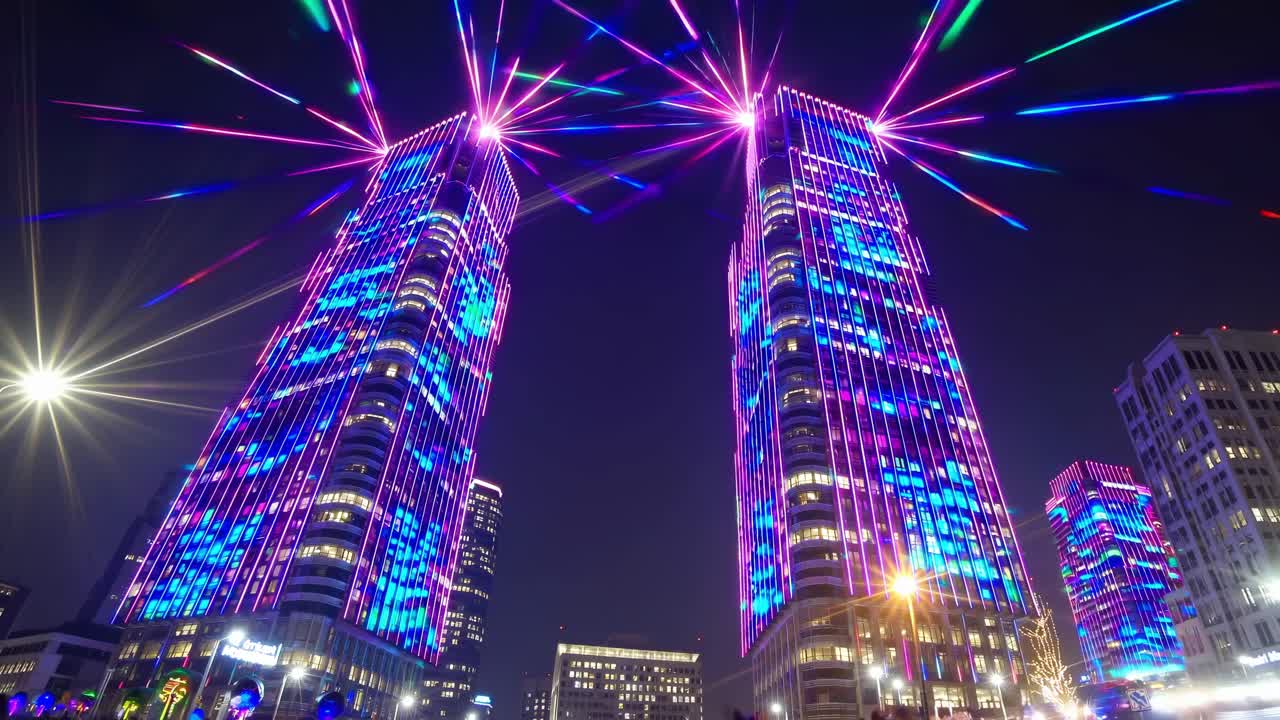 Two modern skyscrapers are displaying a vibrant and colorful light show against the dark night sky, creating a stunning visual spectacle in the heart of a bustling city center