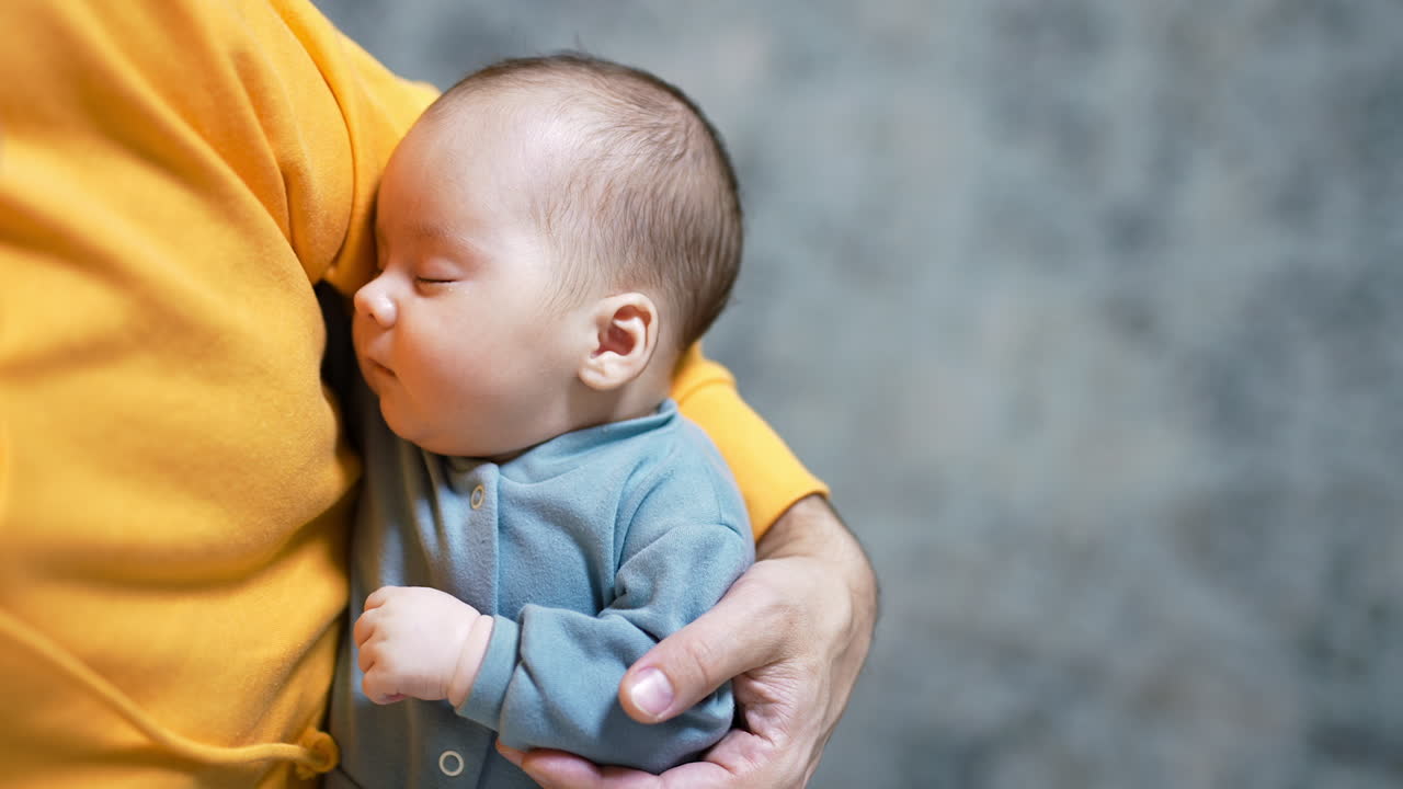 Adorable baby boy in blue clothes resting peacefully in his parent's arms. Male in yellow sweater holding a sleeping baby. Close up.