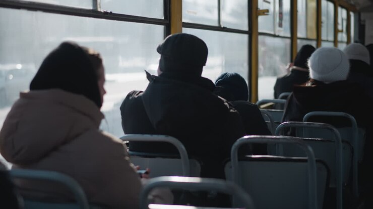 close back view of bundled passengers seated inside moving bus, transparent windows showing bright winter street, soft sunlight and reflections across metal seats create calm urban commute mood