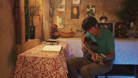 Young-Asian-male-playing-a-guitar-while-sitting-on-a-chair-in-a-rustic-room