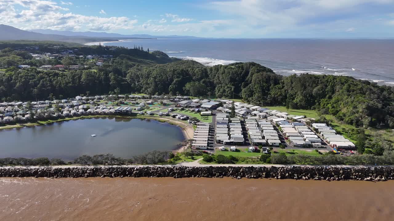 Aerial drone footage over a coastal caravan park beside lagoon and ocean under clear skies