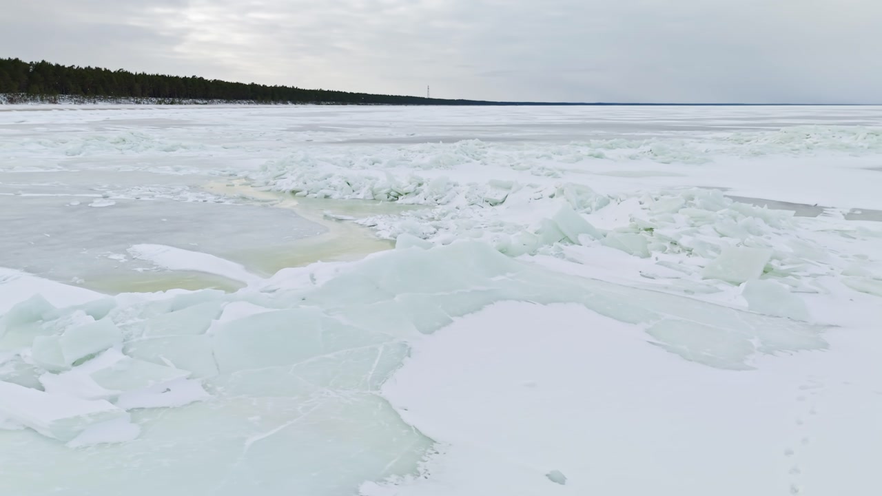 Wide view of frozen lake with pressure ridge, slush pool, and snowy foreground near a dark forest line under cloudy sky. Winter danger and climate thaw concept.