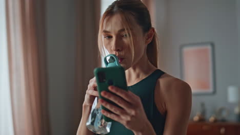 resting sportswoman drinking water looking smartphone at home closeup. slim girl