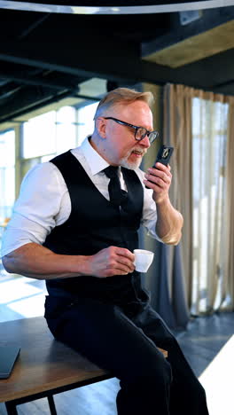 adult male in white shirt, black vest and trousers sits on the desk. man holds a coffee cup in one hand and tries to unblock his phone with nose. vertical video.