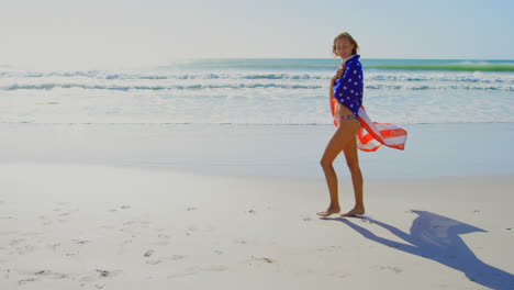 side view of young caucasian woman wrapped in american flag walking on the beach 4k