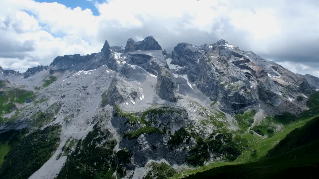 Towering alpine peaks dominate the scene with dramatic ridges, rocky surfaces, and patches of snow, overlooking deep green valleys under a cloudy mountain sky.