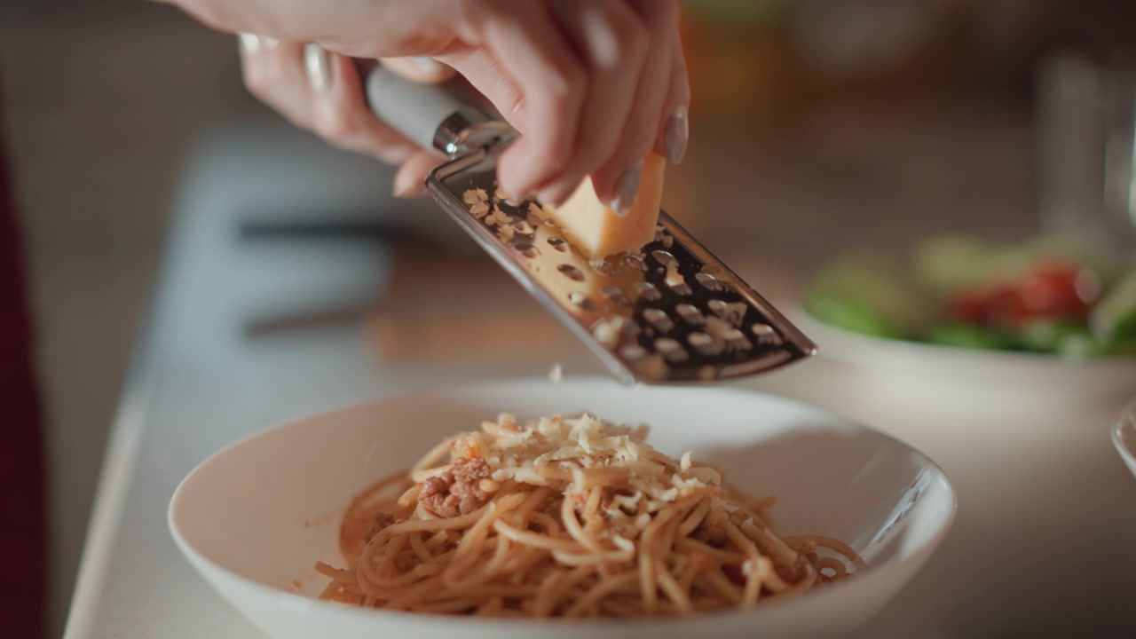 Close-up hand grates hard cheese directly over white shallow bowl of spaghetti, creating downward motion, fresh shavings, and layered topping across noodles in warm home kitchen scene
