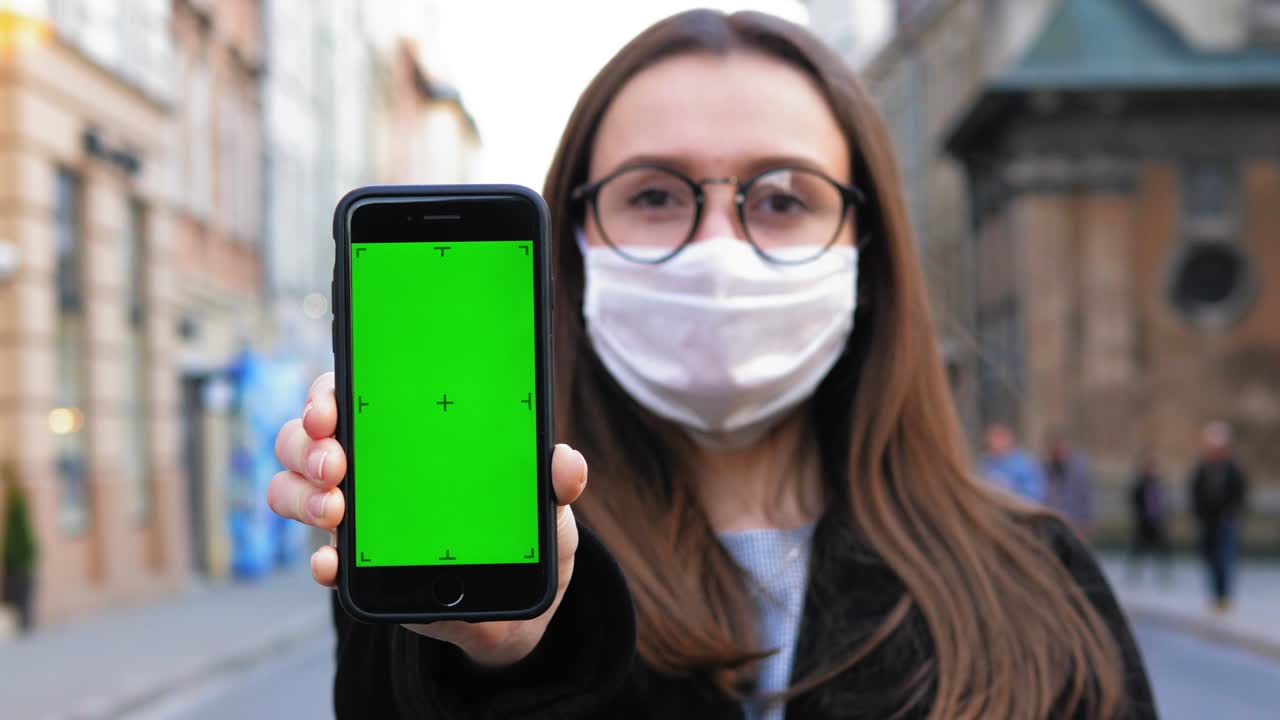 mujer joven de pie con una máscara médica y gafas. sostener el teléfono con pantalla verde. mujer mirando a la cámara. cuidado de la salud y concepto médico de coronavirus. retrato en primer plano en 4k uhd 2160p