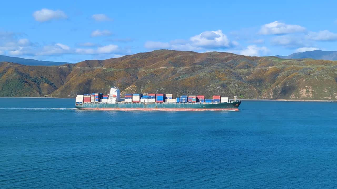 Close up of shipping container ship leaving Wellington harbour in blue ocean water against rugged hill landscape of Wellington, New Zealand Aotearoa