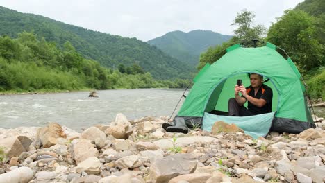 a young tourist sits in a tent on the stone bank of the river and takes pictures on his phone a young tourist sits in a tent on the stone bank of the river and takes pictures on his phone the concept of recreation in nature