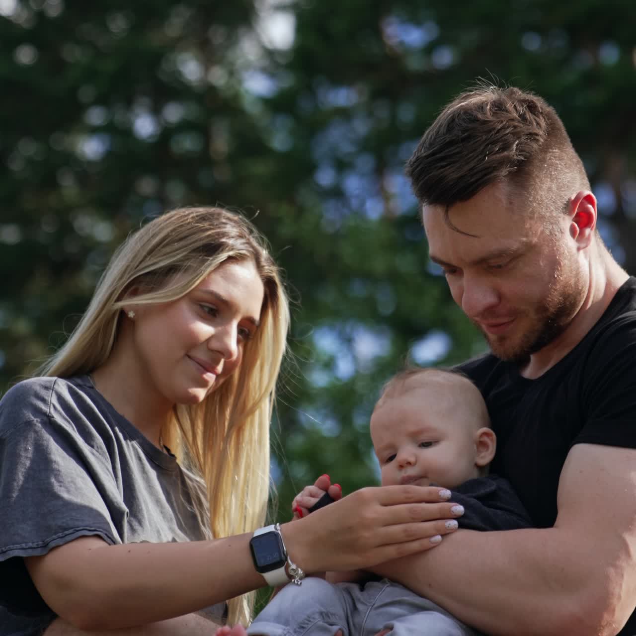 Bearded Caucasian man with a newborn in hands. Woman sitting close stroking child's head with love. Blurred nature at backdrop. Vertical screen