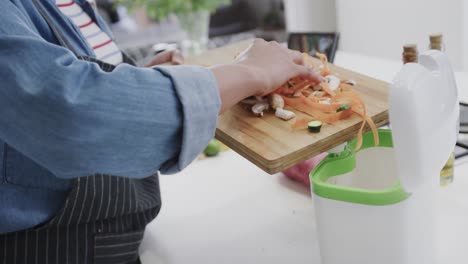 midsection of biracial woman in apron composting vegetable waste in kitchen, slow motion