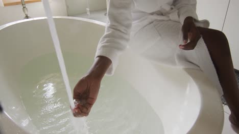 midsection of african american attractive woman in white robe sitting on bathtub, preparing bath