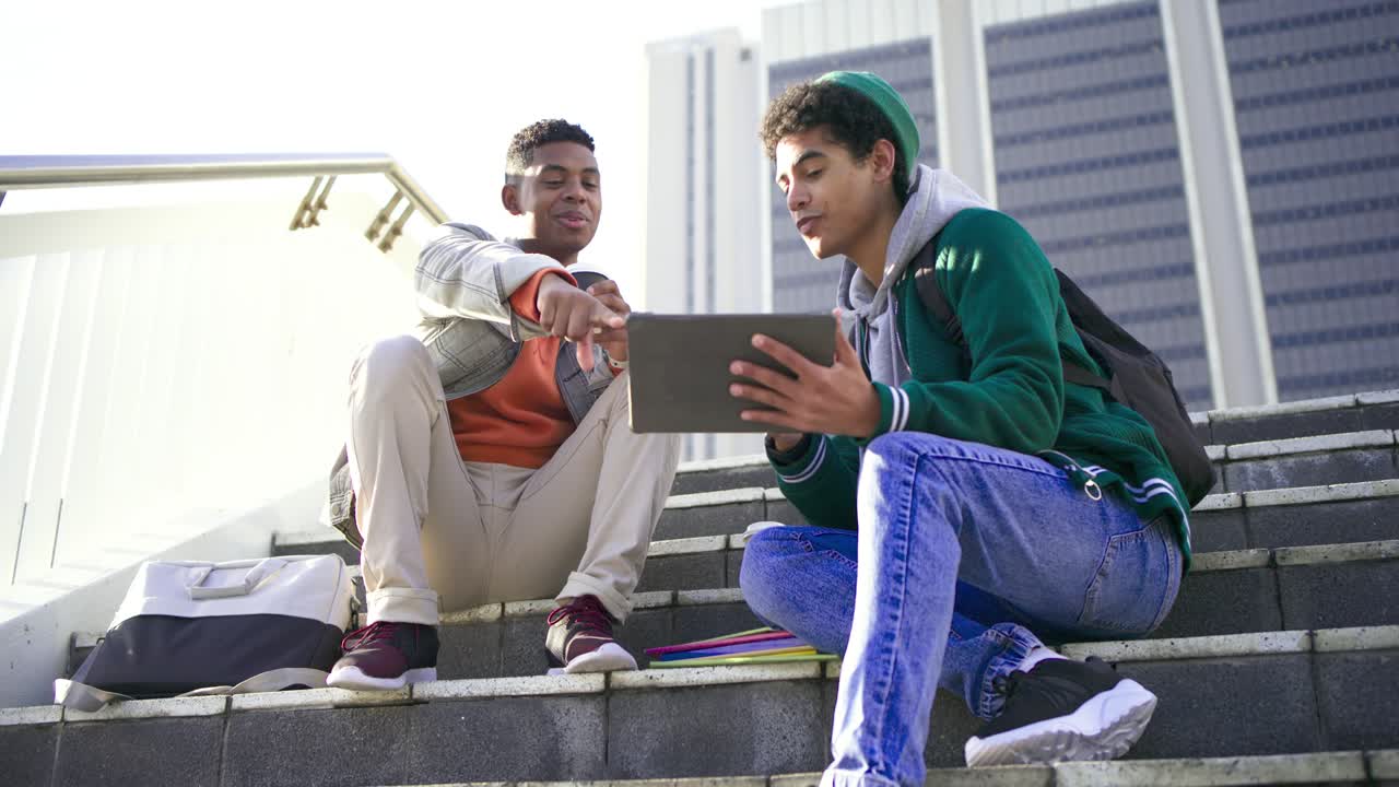 Diverse male friends unfolding tablet, tapping screen, holding coffee and studying on campus steps