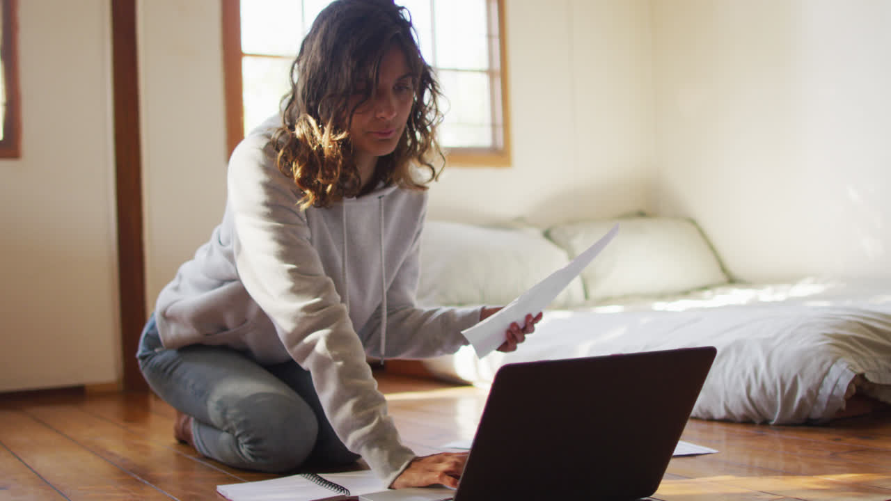 Mixed race woman working at home, kneeling on floor using laptop and holding paperwork in cottage