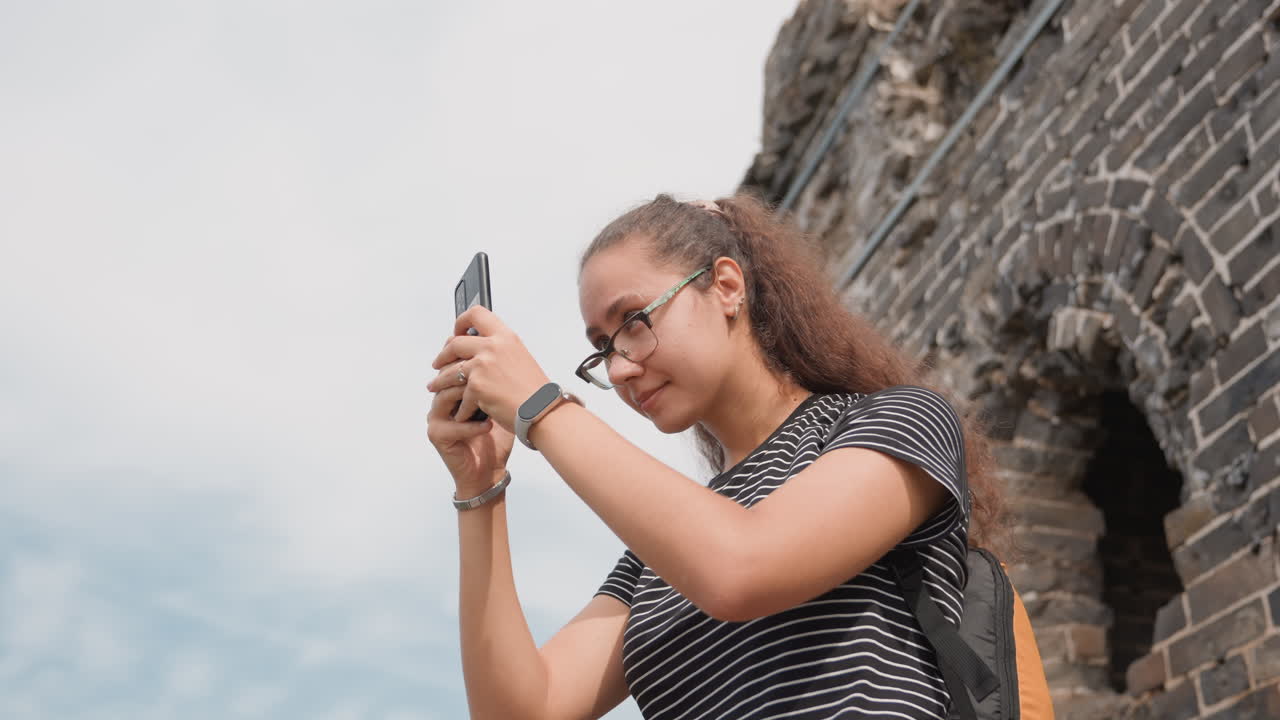 Criadora de conteúdo feminina fotografando ruínas antigas com smartphone, compondo quadros perto de um arco de tijolos desgastados. Carregando uma mochila laranja, usando óculos e camisa listrada. Expressão pensativa.