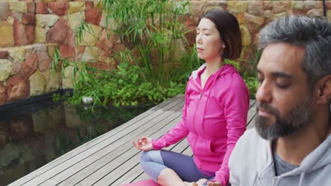 Happy-diverse-couple-doing-yoga,-sitting-and-meditating-at-terrace