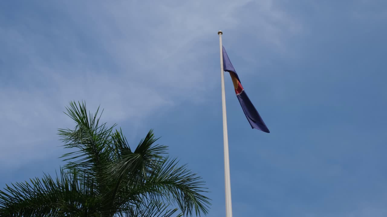 ASEAN Flag flying while the camera tilts a little, a palm tree on the left with a beautiful blue sky and some clouds