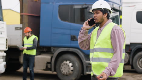 trabajador con chaleco y casco de seguridad organizando una flota de camiones en un parque logístico mientras habla por teléfono