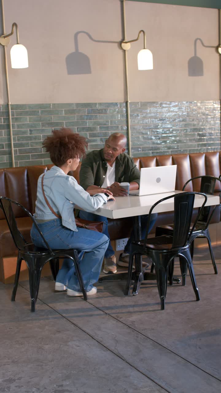 Vertical video: African American team with laptop open discussing while man checking phone at cafe