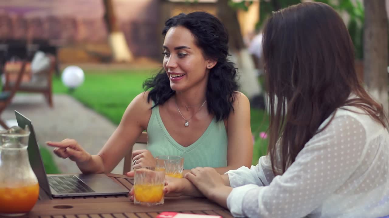 dos mujeres sentadas en el café al aire libre - hablando, compartiendo noticias, chica morena mostrando algo en la pantalla de la computadora portátil y riendo