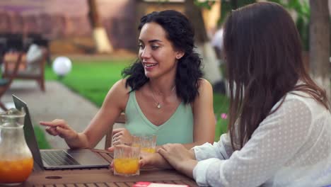 two women sitting at the outdoors cafe - talking, sharing news, brunette girl showing something on laptop screen and laughing