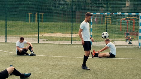 young street football player training freestyle tricks while two team players sitting on the pitch doing stretch exercises and another one using mobile phone 2