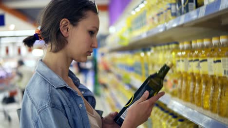 primer plano de una mujer eligiendo productos en un supermercado. leyendo la etiqueta de una botella de aceite. poner una en el carrito. variedad de aceites. filas de botellas con aceite