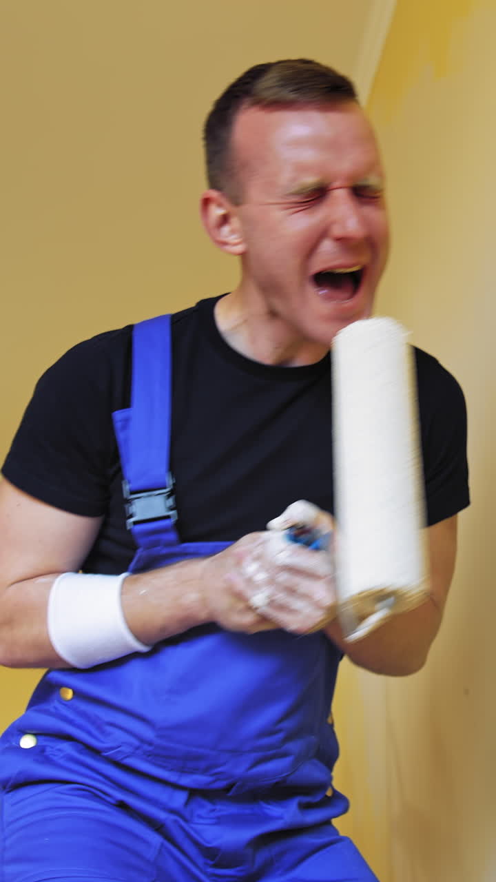 Joyful painter with a paint roller. Funny young man worker in blue overalls singing into a roller while painting walls. Happy moments during makeover in the house. Vertical video