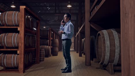 full body side view of an asian male professional worker standing in cellar of winery, he is speaking with someone in the phone and moving his left hand