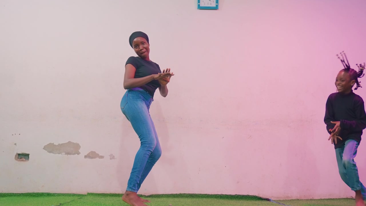 Madre e figlia di colore che ballano all'aperto contro un muro chiaro sotto una luce rosa, indossando jeans sull'erba verde, coreografia giocosa, legame gioioso, sorrisi espressivi e ritmo condiviso.