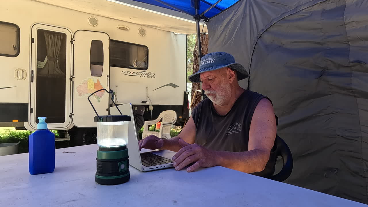 En person arbejder på en bærbar computer på en campingplads. Omgivelserne inkluderer en autocamper og udendørsudstyr. Aktiviteten er afslappet og viser balancen mellem arbejde og natur i et campingmiljø.