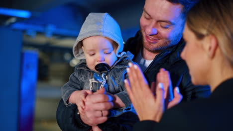 dad holds a baby looking focused at man's hands. mother tries to attract child's attention clapping hands.