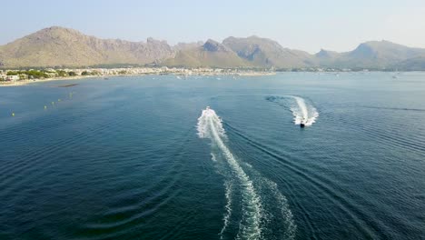 Drone-shot-of-two-speed-boats-cruising-at-a-high-speed-in-the-waters-of-the-Mediterranean-Sea,-off-the-coast-of-Playa-del-Port-de-Pollenca-beach-resort-in-Mallorca,-Spain