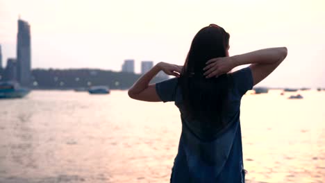 young woman in summer dress standing on the beach of tropical city and looking to a sea at sunset