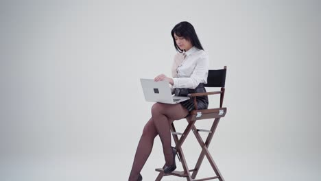 Sexy young lady open laptop in studio. Beautiful woman in white blouse and short skirt sitting on a chair and holding laptop on light background. Slow motion.