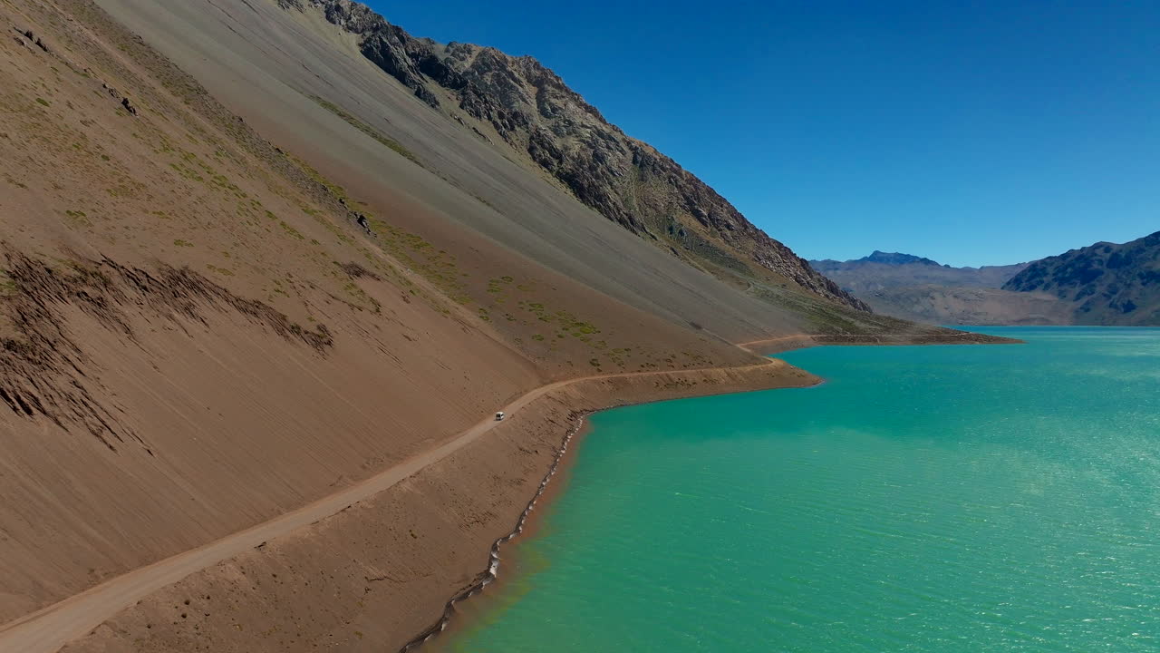 Car driving on dirt road alongside turquoise water of Embalse El Yeso reservoir, rugged Andes mountains under clear blue sky, Chile. Aerial forward