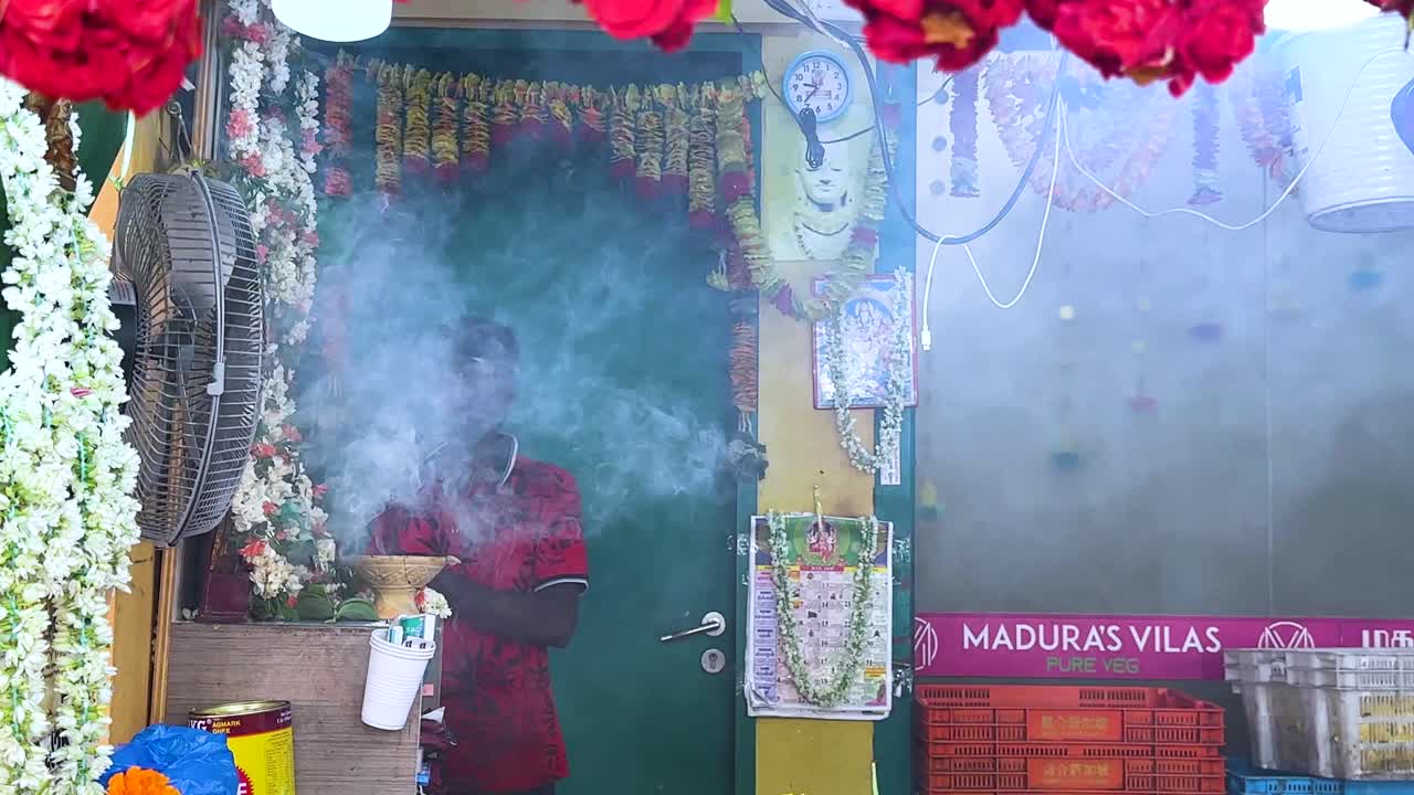 A vendor tends incense amid vibrant jasmine and rose garlands at a colorful Singapore market stall. Daylight, static camera, close-up and wide shots