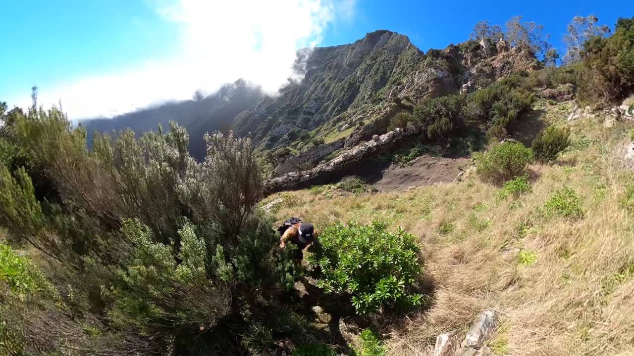 A young and strong solo man hiker is holding a selfie stick with a 360 action camera as he walks up the mountainside of Espigao Amorelo mountain in Madeira with the harch sun during a clear day.