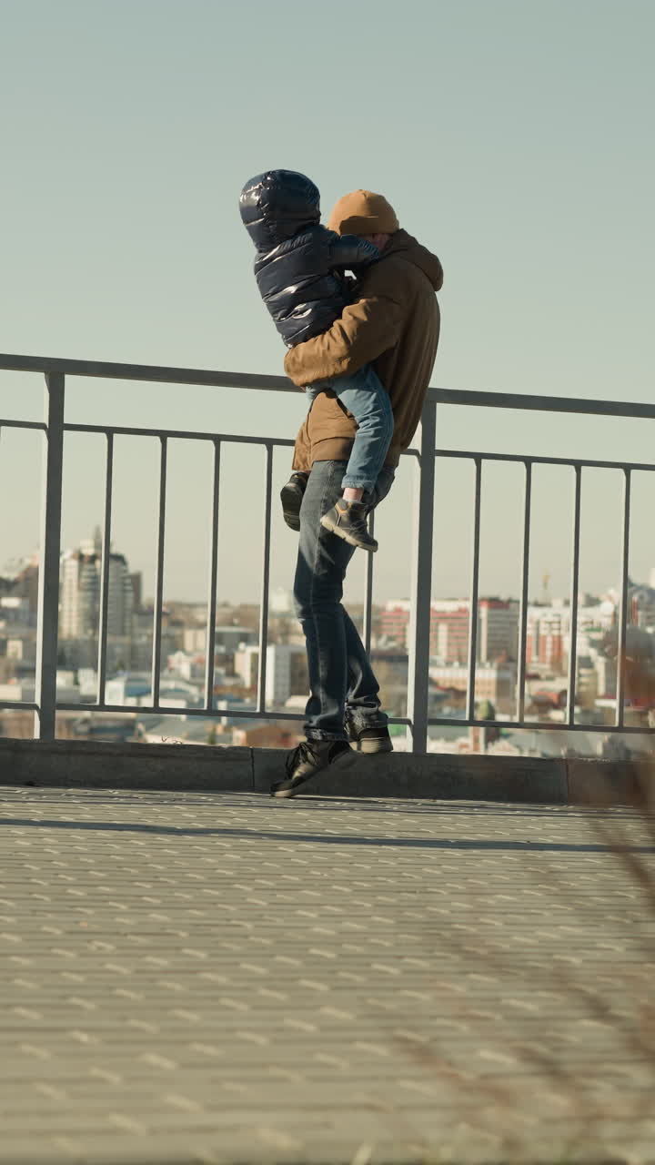 A father tenderly holds his son close to an iron railing, stepping down and then back up, both observing the cityscape below, they are both dressed jacket and beanie