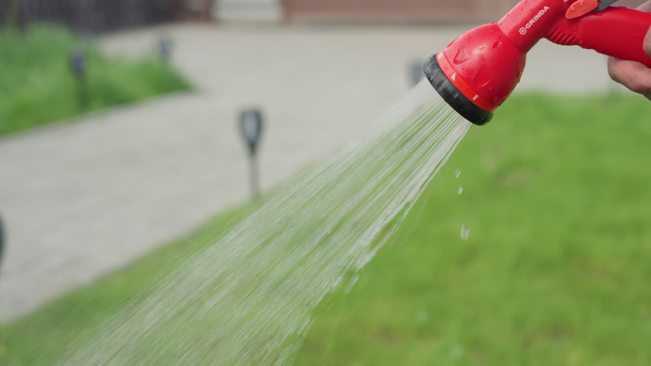 close up of hand spraying water over green garden grass through red nozzle, droplets dancing in daylight capturing nurturing care routine, soil and blades glistening under radiant sun