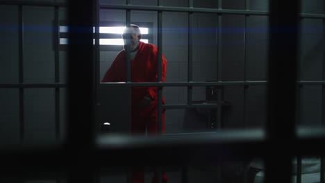 elderly prisoner in orange uniform holds hands on metal bars