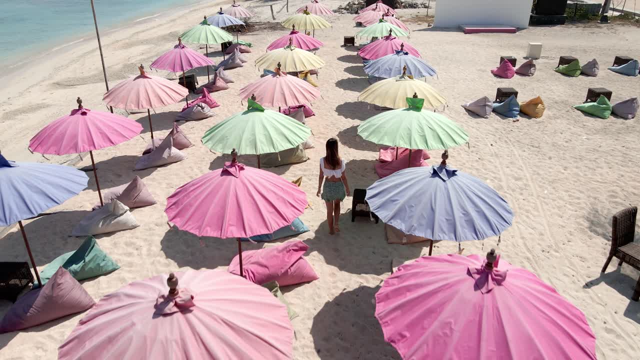 다채로운 우산 사이의 모래 해변을 걷는 여성의 공중 슬로우 모션 샷